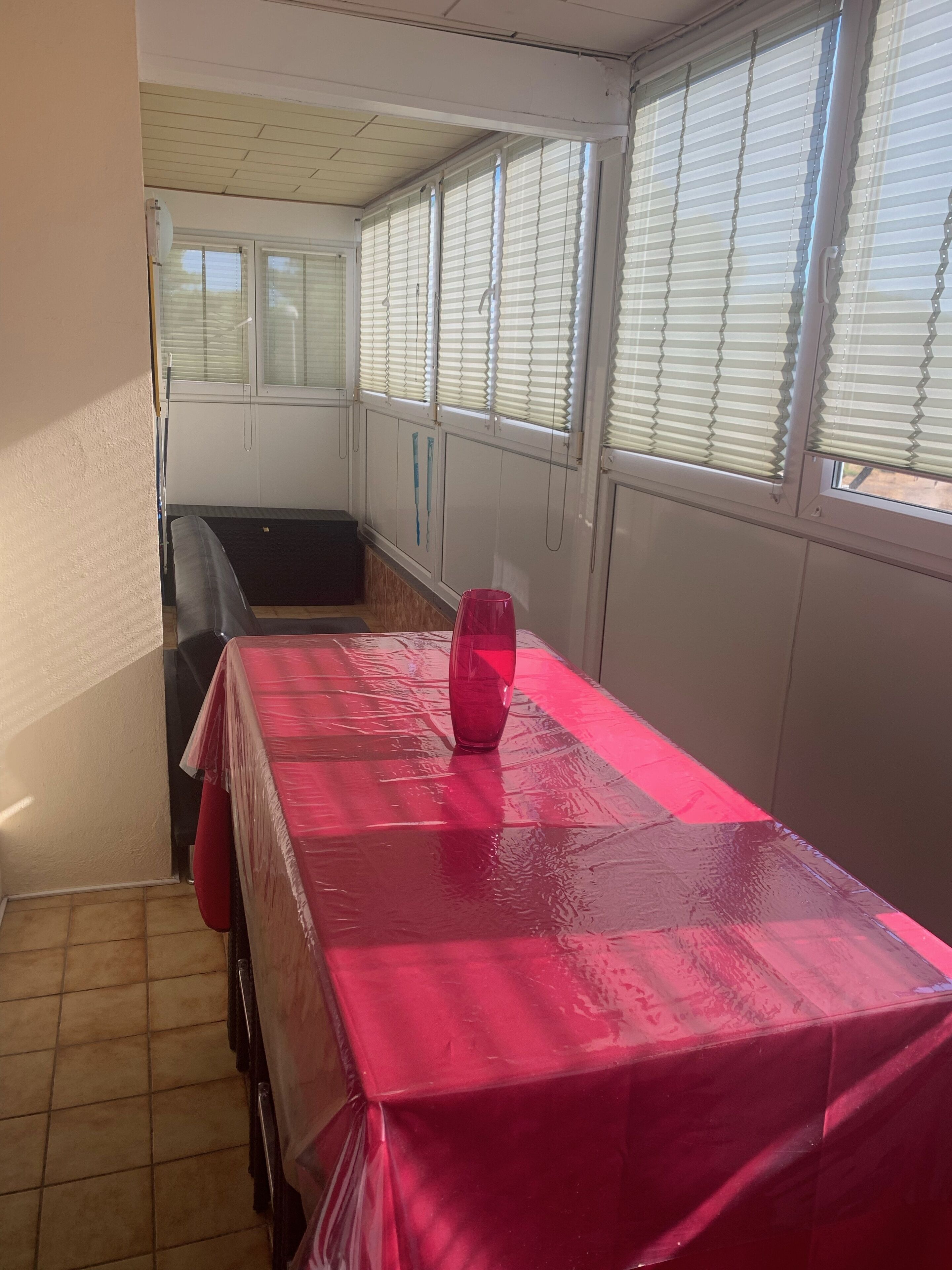 Une pièce avec un sol carrelé, une table recouverte d’une nappe rouge, un vase rouge et une fenêtre avec des stores.