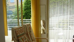 A room with two rocking chairs, a table, and a view of a balcony with yellow curtains.