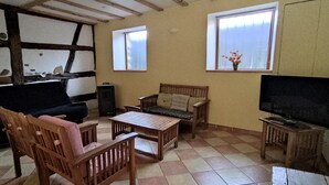 Un salon avec un canapé, des chaises en bois, une table basse, un téléviseur et deux fenêtres.