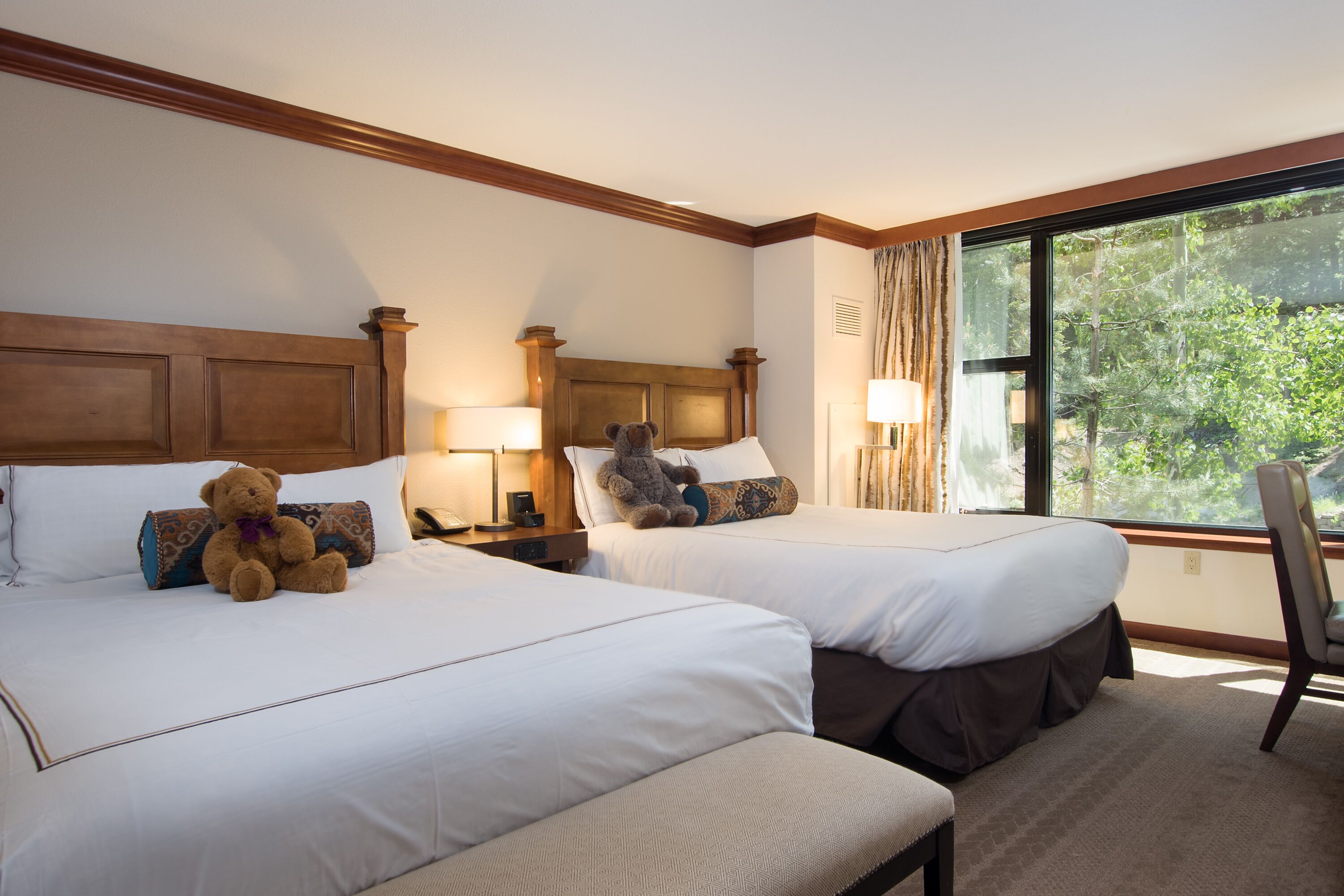 A hotel room with two beds, each with a headboard, a large window with a view of trees, and a teddy bear on one bed.