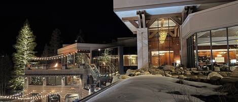A modern building with a glass facade, outdoor seating, and a snow-covered slope.