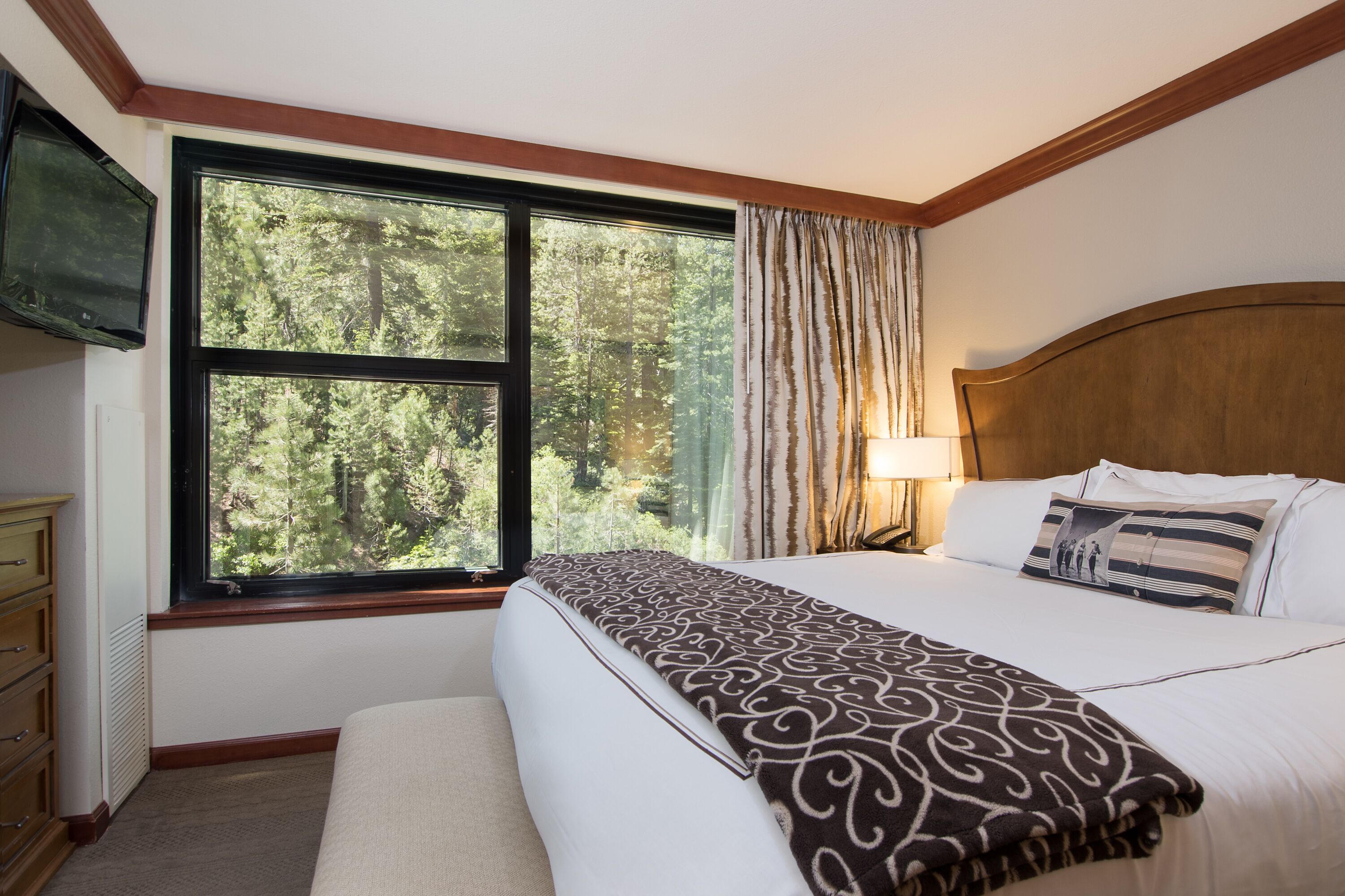 A hotel room with a large window, a bed with a patterned blanket, a bedside lamp, and a television on the wall.