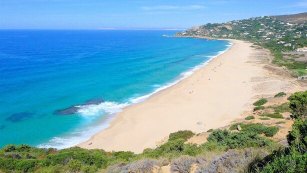 A coastal landscape with a sandy beach, clear blue sea, and a hilly area with vegetation.