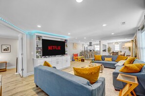 A modern living room with a flat-screen TV displaying 'NETFLIX', a sofa set, and a dining area with a table and chairs. - The Grand Ranch: 6-BR Luxury Retreat with with Gazebo, Fire Pit & Outdoor Cinema (Raleigh)