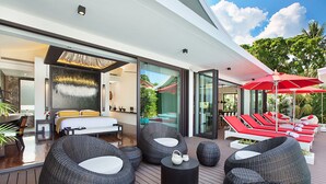 Un espace de détente extérieur moderne, avec des meubles en osier, un parasol rouge et une vue sur la piscine et la verdure.