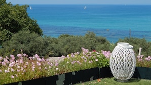 Un jardín con una lámpara decorativa blanca, diversas flores y vista al mar con veleros.
