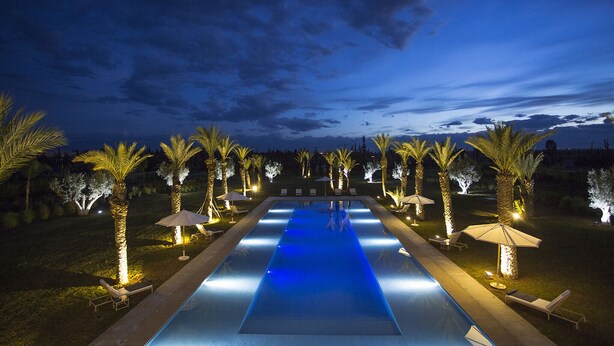 A swimming pool with a blue-lit water feature, surrounded by palm trees and lounge chairs. - ADNAA - Luxury Villa with 2 pools, sauna, hammam, tennis court & home cinema (Marrakech)
