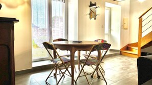 A dining area with a round table and four chairs, a window with curtains, a mirror on the wall, and a staircase.