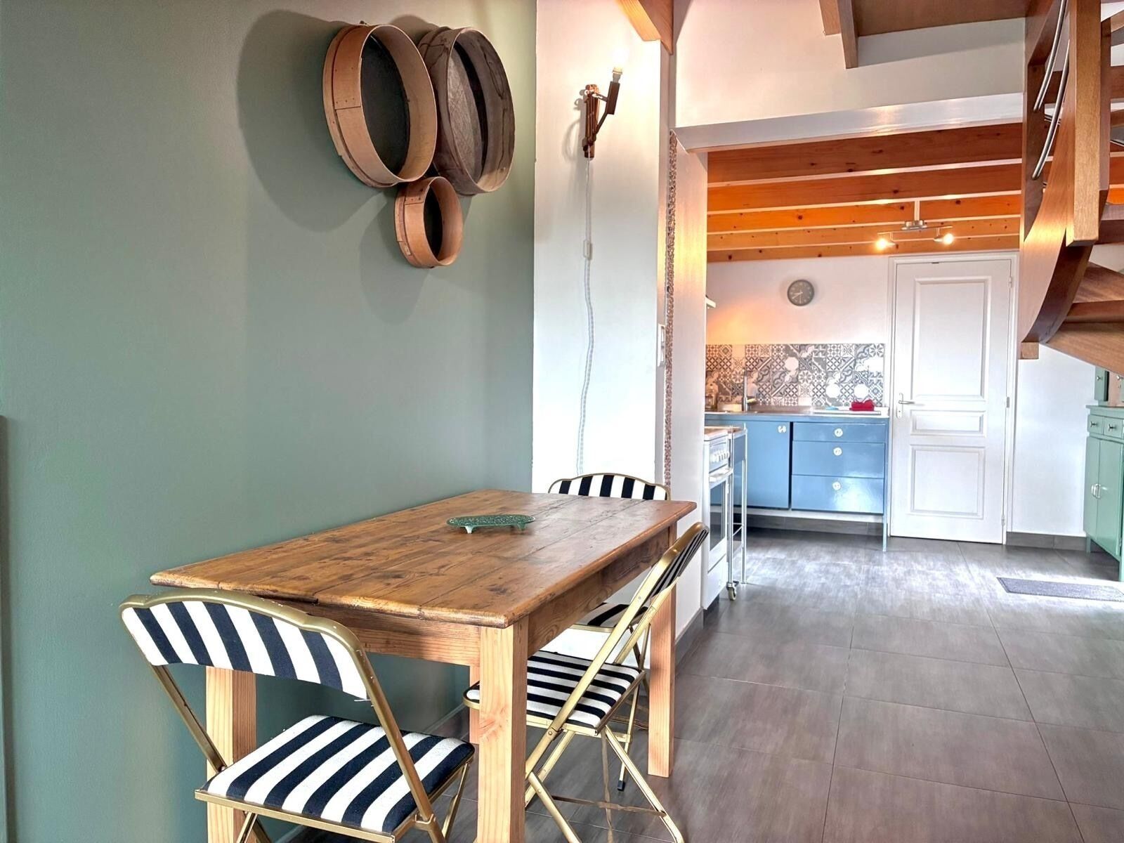 Un coin repas avec une table en bois et des chaises à rayures, une cuisine en arrière-plan et un escalier avec des poutres en bois.