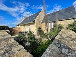 A stone house with a sloped roof, a stone wall, and a garden with various plants.