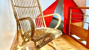 A wicker chair with a high backrest and armrests, positioned in a corner of a room with a wooden floor and a red and beige geometric wall design. - Reder Mor : maison de pêcheurs 250 m de la mer (SANTEC)