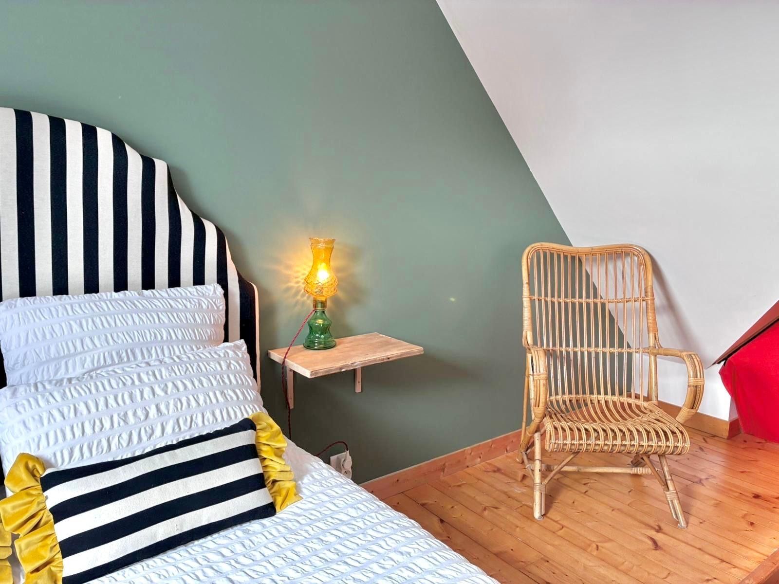 A bedroom with a bed, a nightstand, a lamp, and a wicker chair.