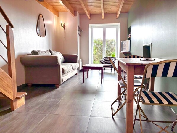 A modern living room with a sofa, a dining table with chairs, a TV, and a large window with a view of greenery.