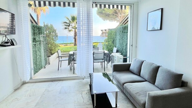Un salon moderne avec un canapé gris, une table basse en verre et un coin repas comprenant des chaises et une table. Une grande fenêtre offre une vue sur la mer et la verdure.