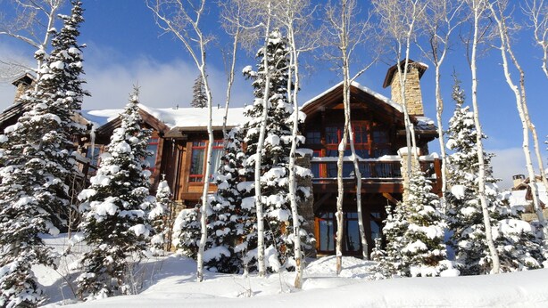 Un chalet enneigé, doté d’une cheminée en pierre, entouré d’arbres chargés de neige.
