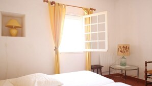 Ein Zimmer mit einem Bett, einem Fenster mit gelben Vorhängen, einem kleinen Tisch mit einer Lampe und einem Stuhl.