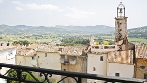 Blick auf eine Stadt mit Häusern mit Terrakotta-Dächern, einem Glockenturm und sanften Hügeln im Hintergrund.