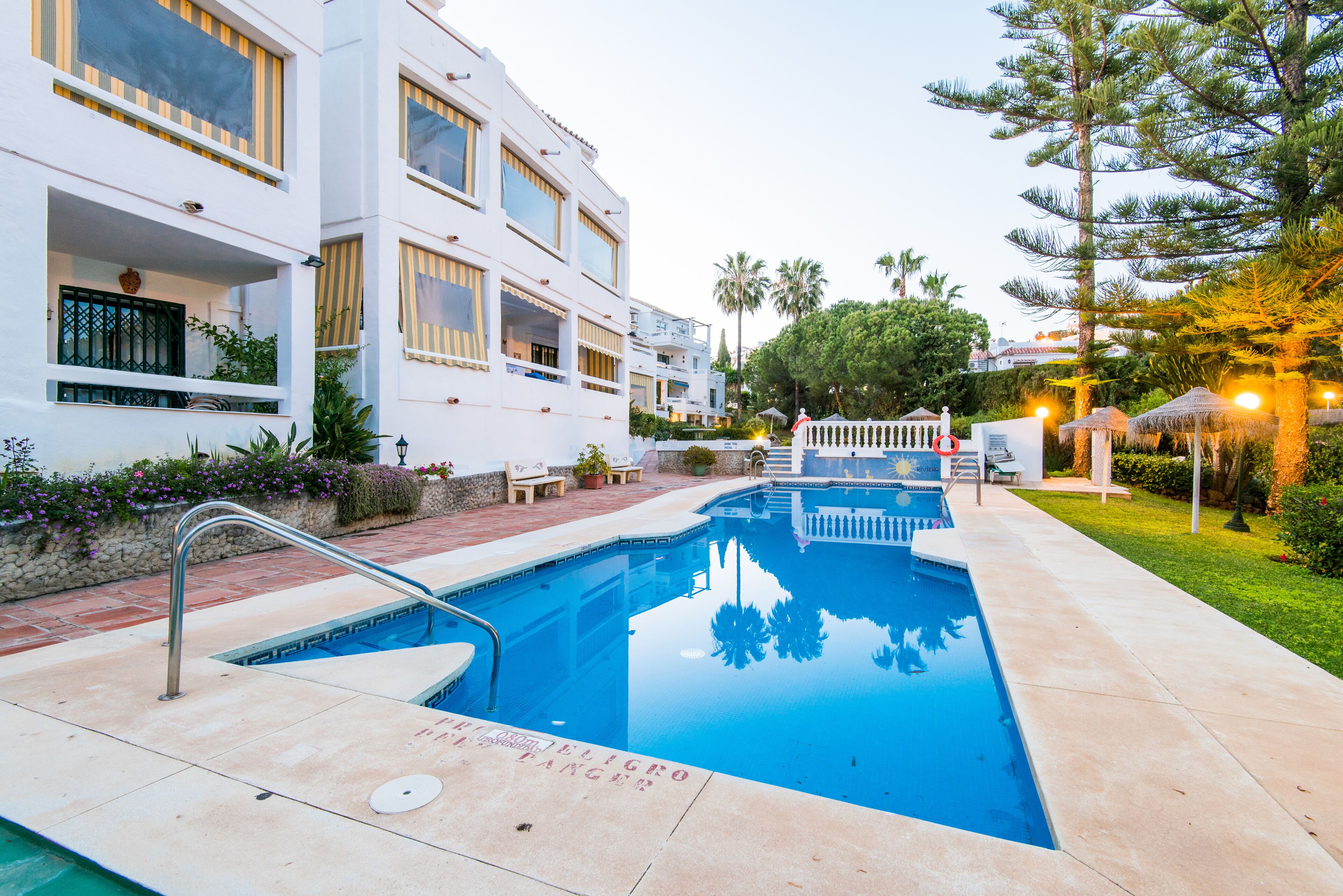 A swimming pool with a metal handrail, surrounded by a paved area with a white fence and a bench.