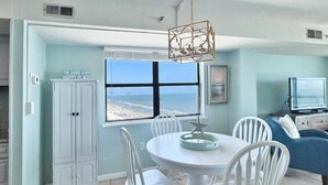 Een eetruimte met een ronde tafel, witte stoelen, uitzicht op het strand, een kast en een televisie.
