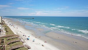 Een strand met een steiger die in de oceaan uitloopt, een zandstrand met mensen en parasols, en een gebouw met een trap die naar het strand leidt.