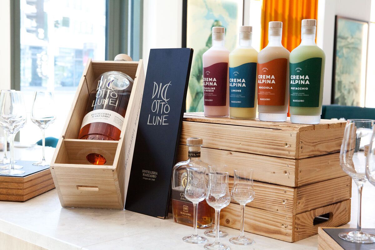 Eine Auswahl an Crema Alpina Likören in Holzkisten, zusammen mit einer schwarzen Schachtel und einer Flasche Distilleria Marzadoro.