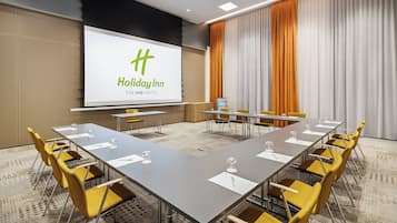 Ein Konferenzraum mit einer großen Leinwand, auf der das Holiday Inn Logo zu sehen ist, einem langen Tisch mit Stühlen und einem Teppichboden.