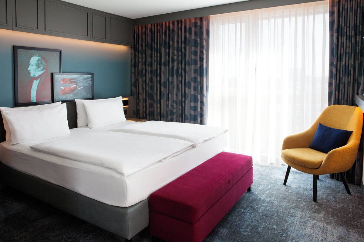 A double bed with white linens, a red ottoman, a yellow armchair, and a large window with sheer curtains.