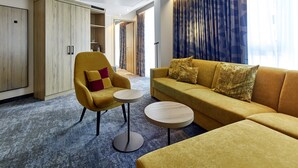 A modern hotel room with a yellow sofa, a matching armchair, and a small round table. There is a wardrobe and a door leading to another room. - Holiday Inn Hamburg - Hafencity by IHG (Hamburg)