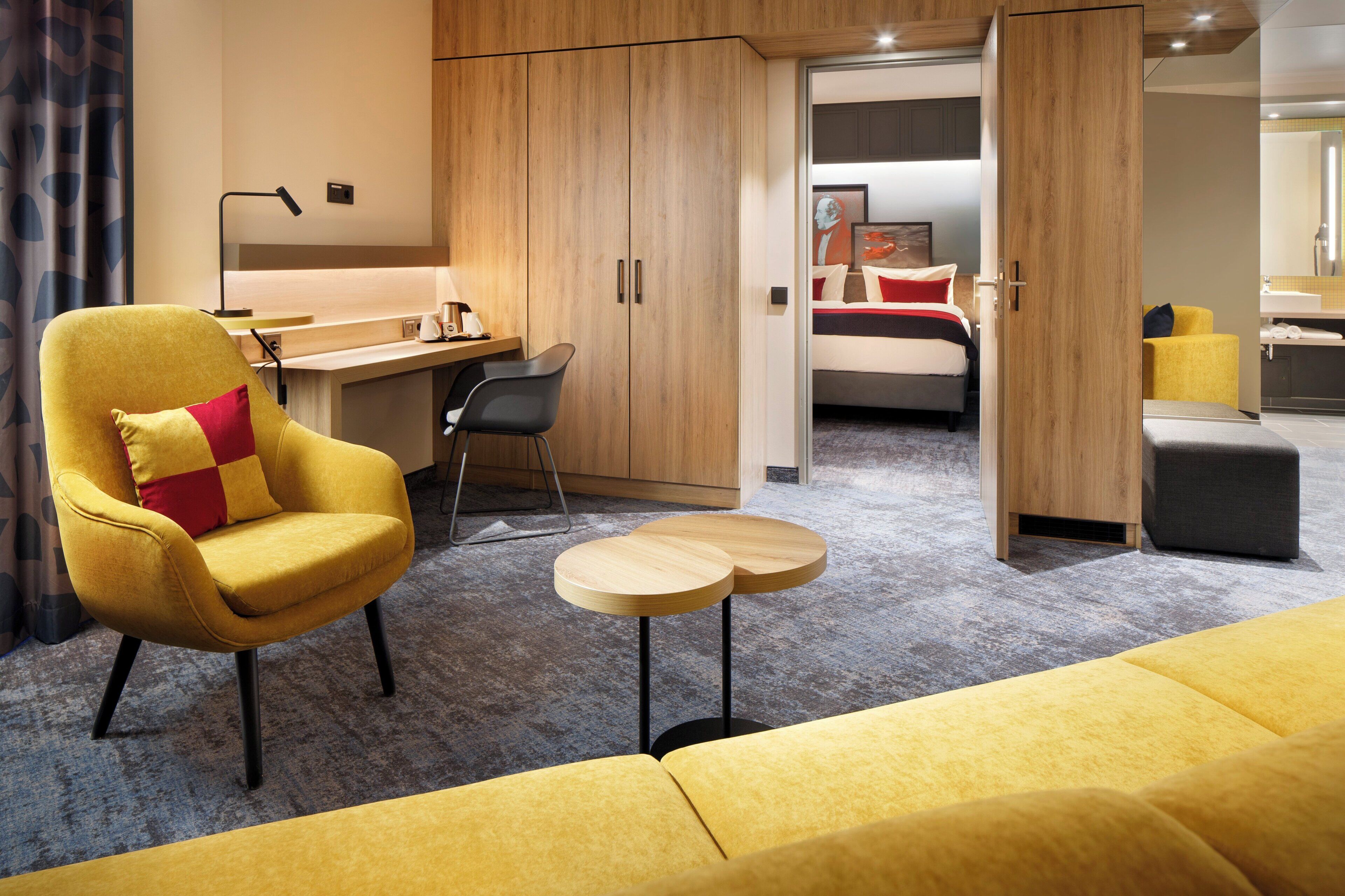 A modern hotel room with a yellow armchair, a small round table, a bed with red and white bedding, and a bathroom visible through an open door.