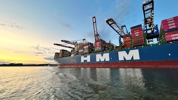 Un gran buque de carga con las letras "HMM" en su lateral, atracado en un puerto donde grúas cargan y descargan contenedores.