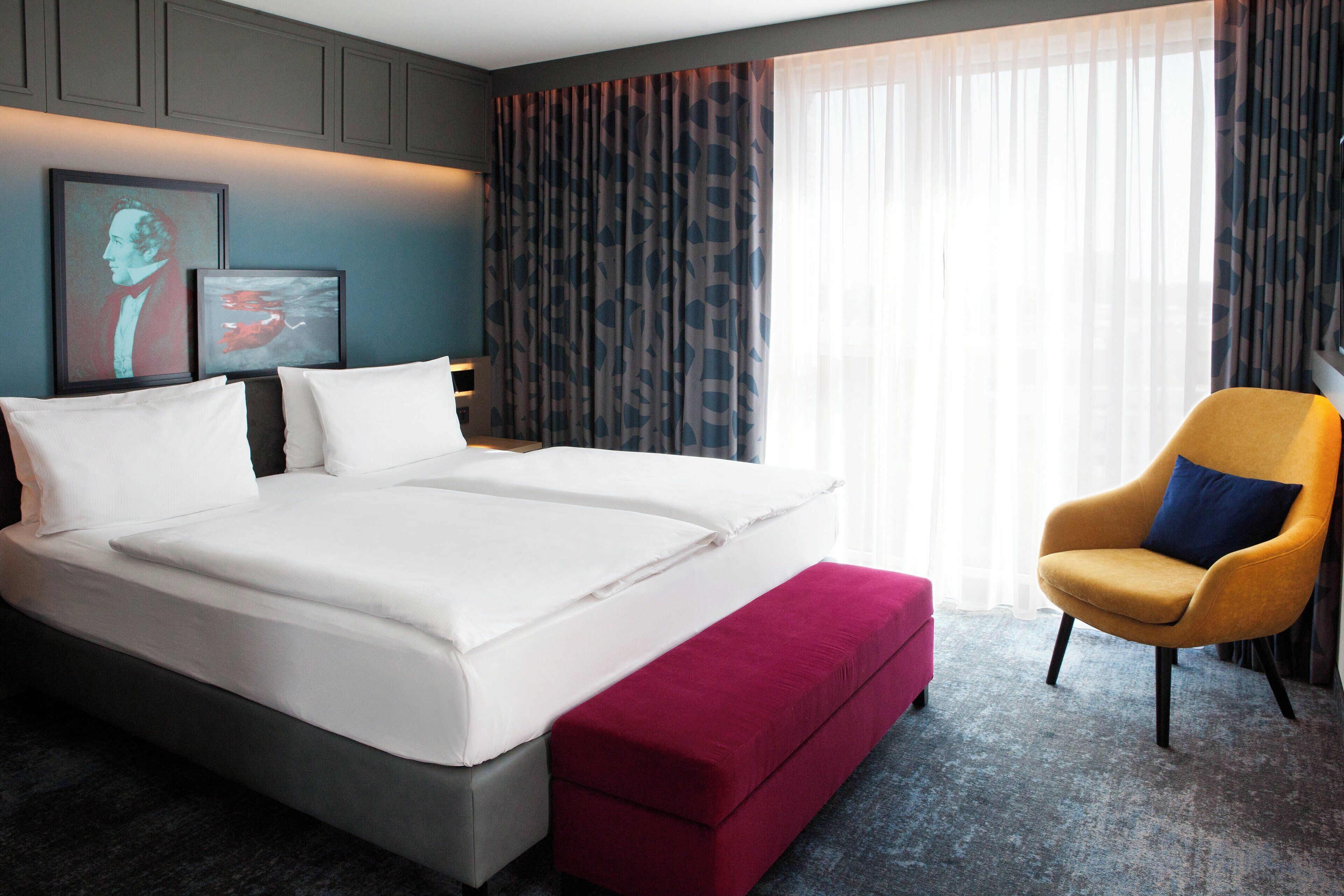 A hotel room with a large bed, a red ottoman, a yellow armchair, and a large window with curtains.