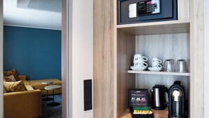A hotel room with a wooden shelf containing a coffee maker, a kettle, and a box of coffee pods. There is a sofa and a small table in the background. - Holiday Inn Hamburg - Hafencity by IHG (Hamburg)