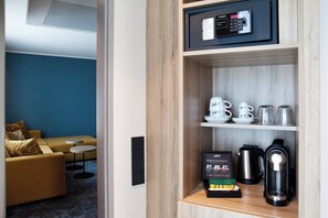 A hotel room with a wooden shelf containing a coffee maker, a kettle, and a box of coffee pods. There is a sofa and a small table in the background. - Holiday Inn Hamburg - Hafencity by IHG (Hamburg)