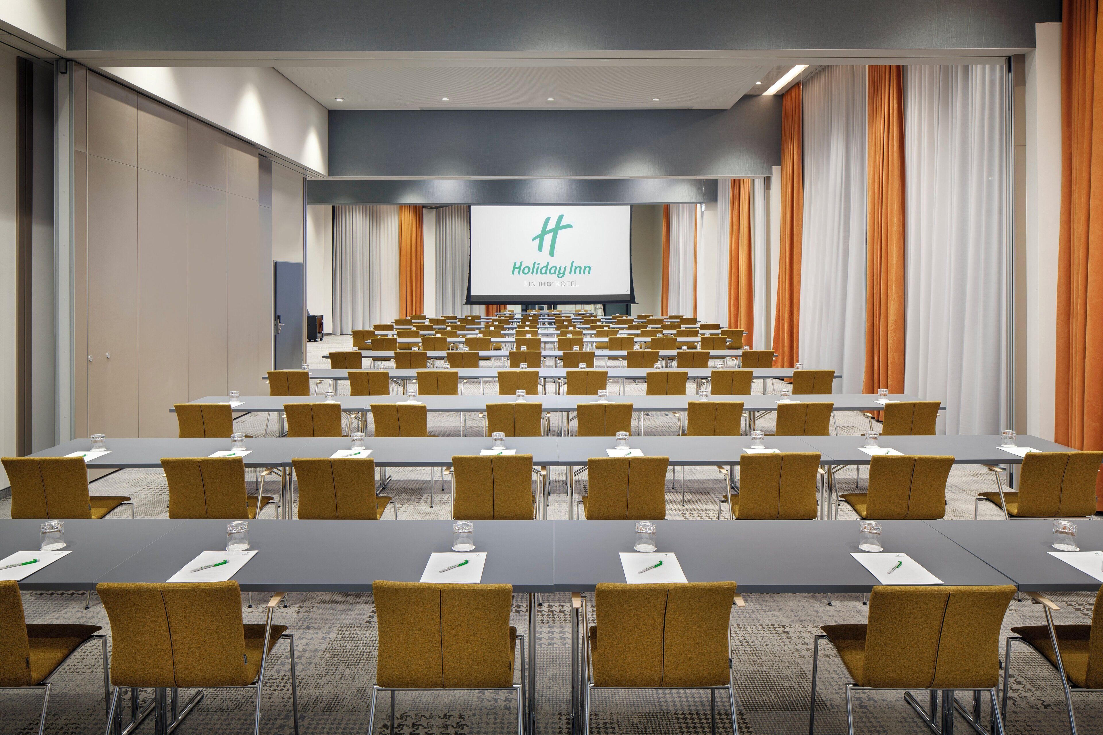 Une salle de conférence avec des rangées de tables et de chaises, un grand écran à l’avant et le logo de l’hôtel Holiday Inn.