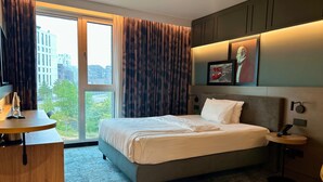 A hotel room with a large bed, a desk with a lamp, a small table, and a view of buildings and trees through the window. - Holiday Inn Hamburg - Hafencity by IHG (Hamburg)