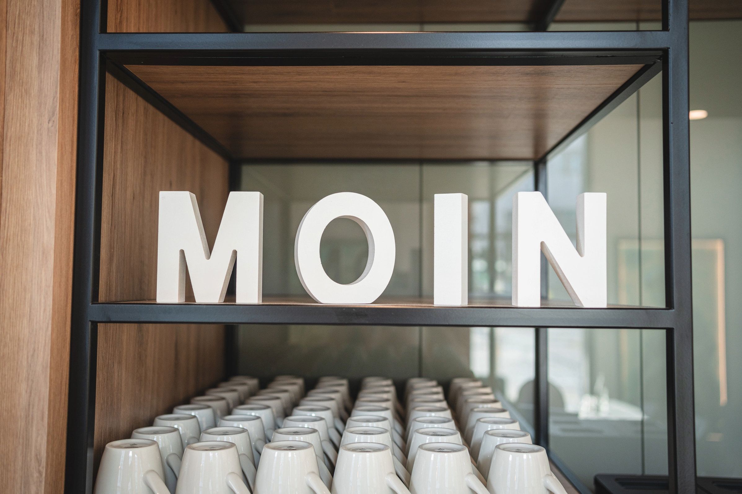 A display of white mugs in a wooden and black-framed shelf with the letters "MOIN" above them.