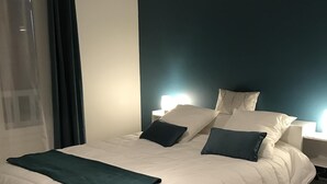 Un lit bien fait, avec du linge de lit blanc et un plaid turquoise, un tapis blanc et moelleux, et un mur d’accent bleu foncé.