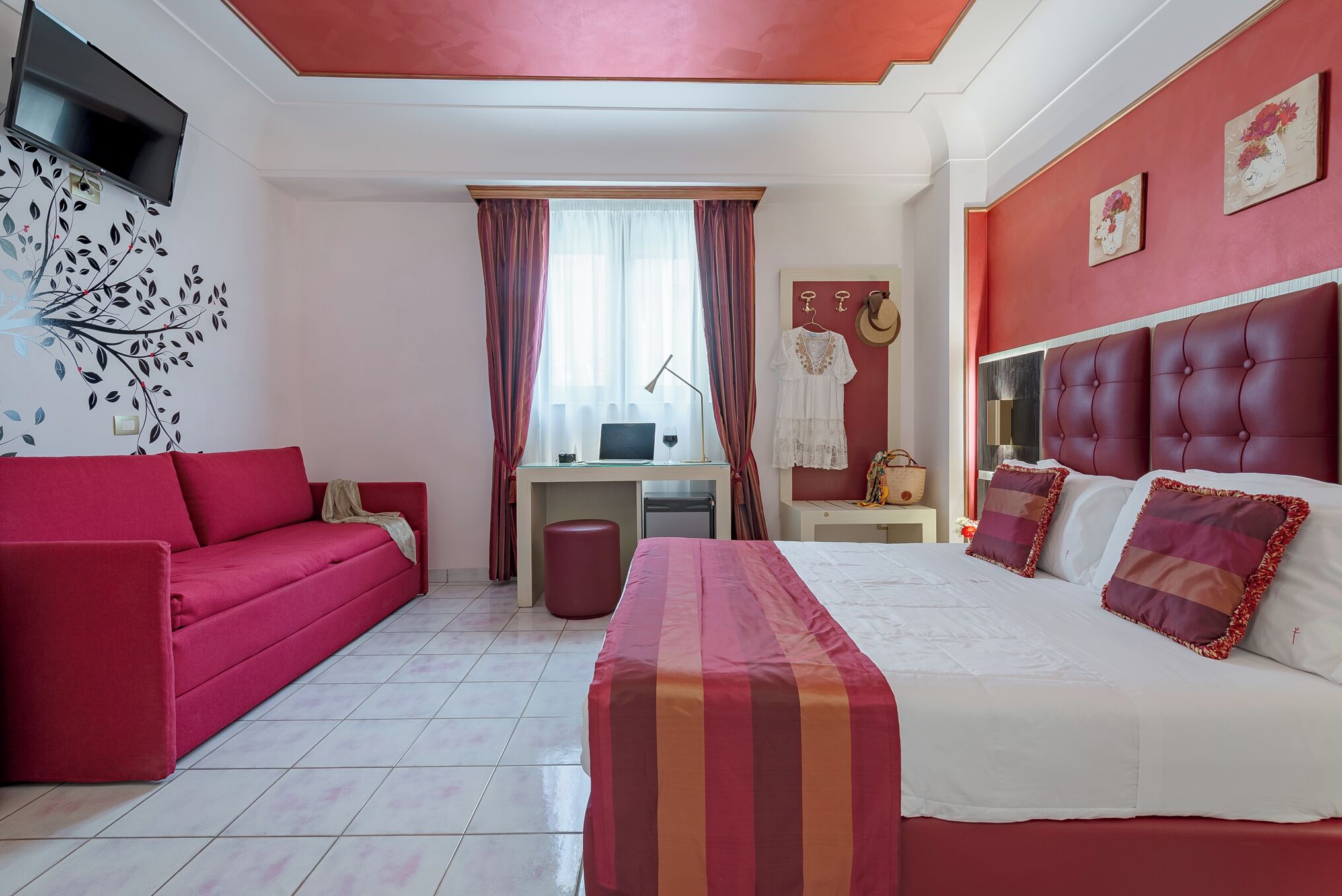 A room with a red sofa, a bed with striped bedding, a desk with a computer, and a TV mounted on the wall.