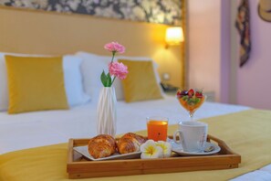 Una bandeja de desayuno con croissants, un vaso de jugo de naranja, una taza de café y un cuenco de frutas sobre una cama con almohadas y un reloj de pared.