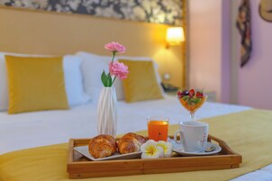 A breakfast tray with croissants, a glass of orange juice, a cup of coffee, and a fruit bowl on a bed with pillows and a wall clock.