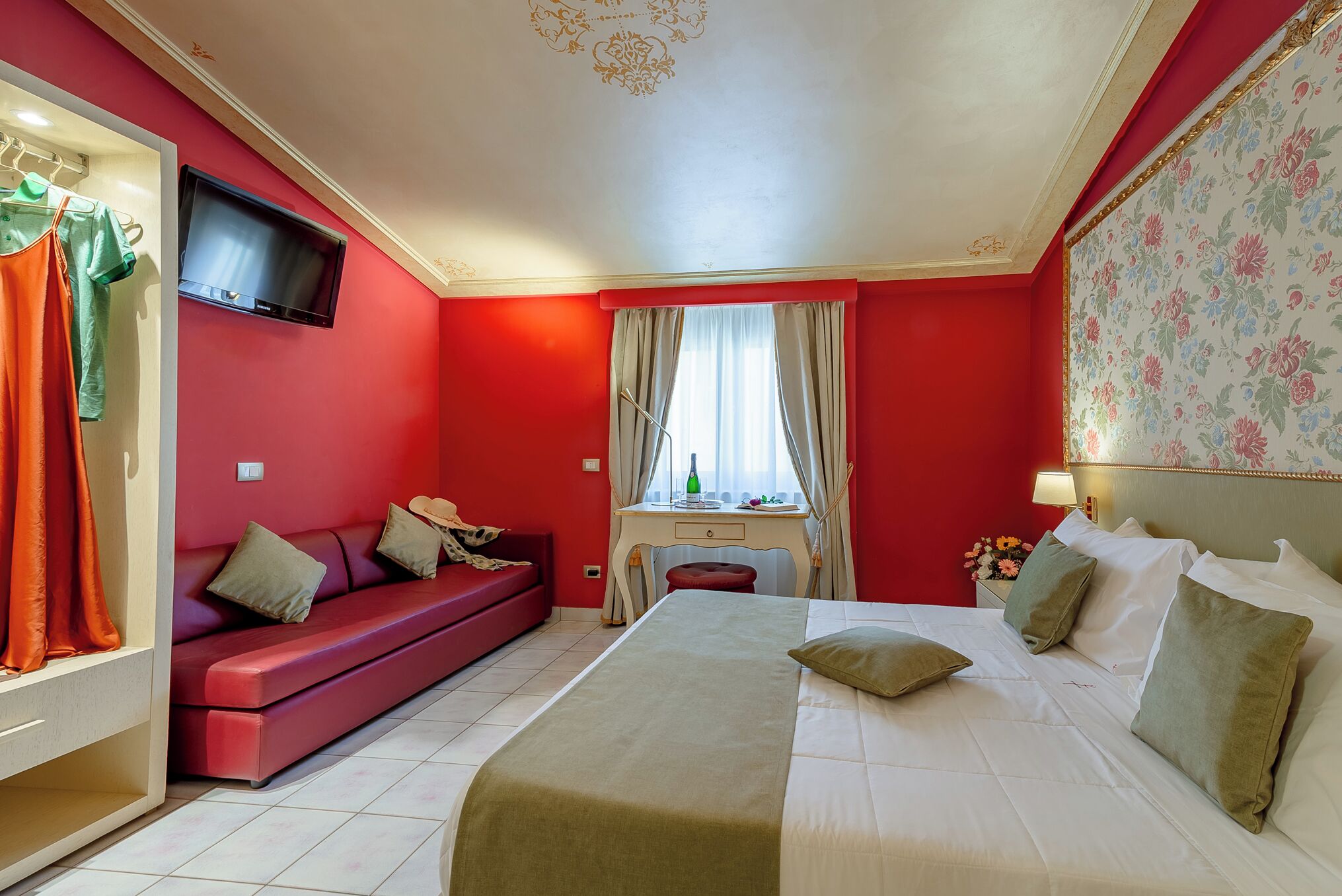 Habitación de hotel con pared roja, cama con ropa blanca, sofá con cojines, televisor montado en la pared, escritorio con lámpara y ventana con cortinas.