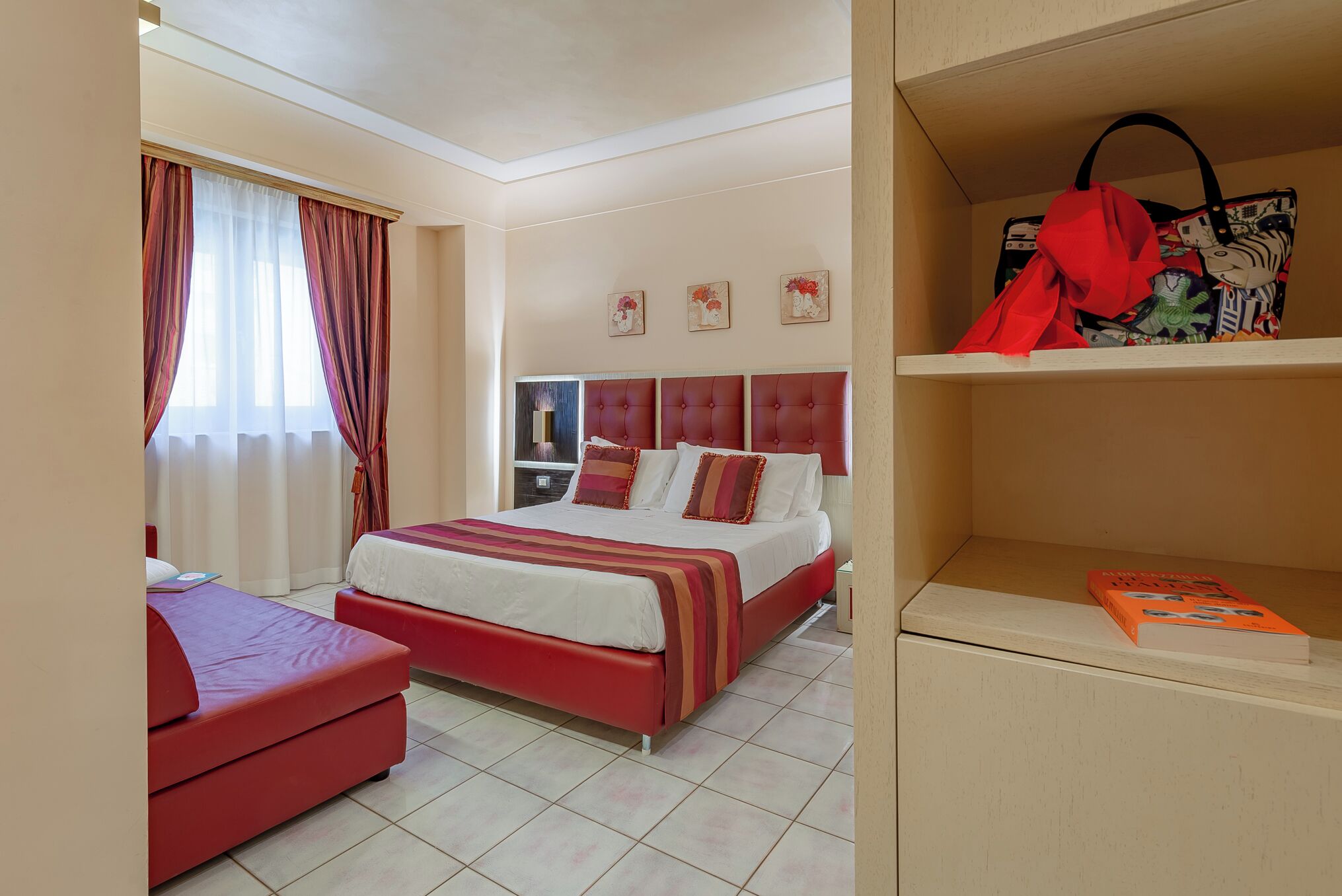 A hotel room with a bed, a red sofa, a bookshelf, and a window with curtains.
