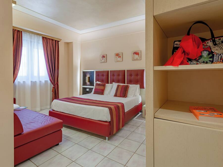 Una habitación de hotel con una cama, un sofá rojo, una estantería y una ventana con cortinas.