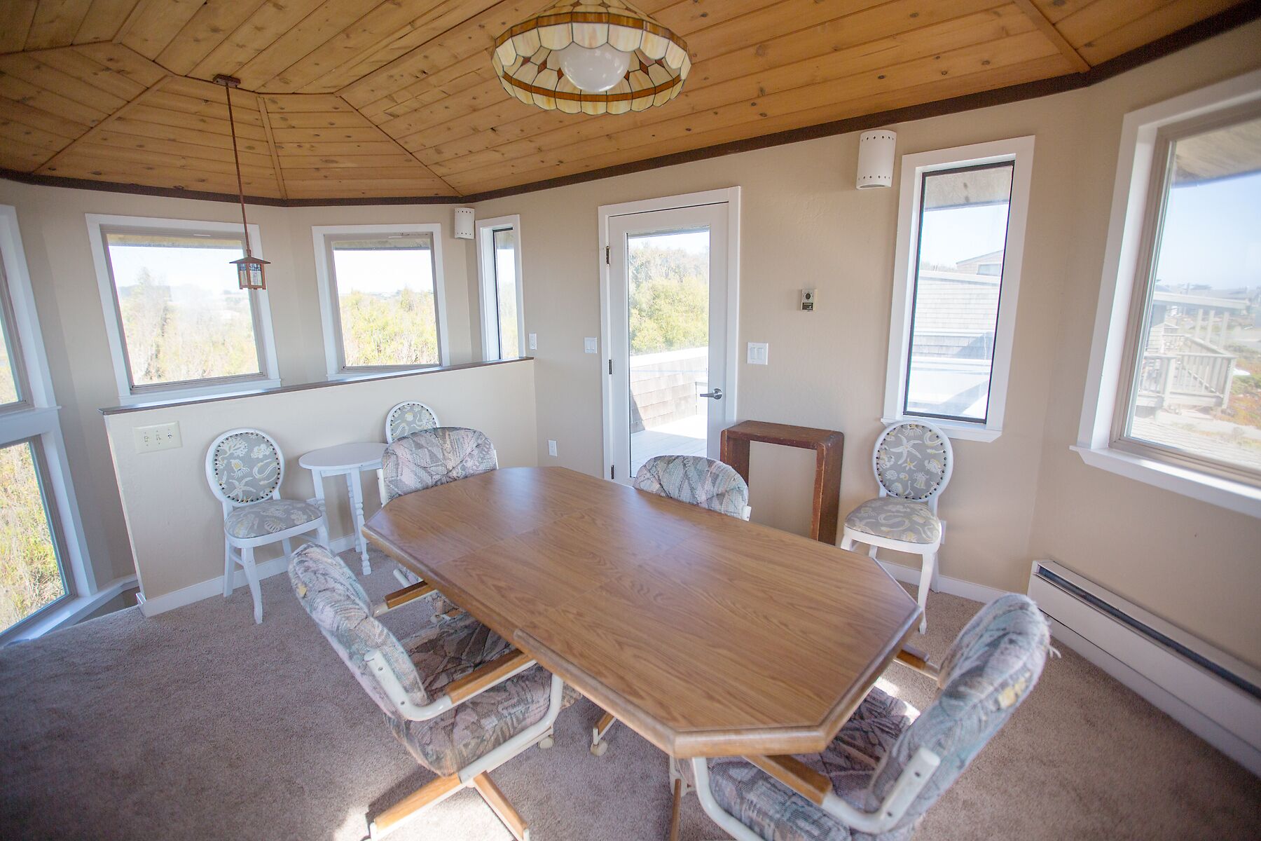 A spacious room with a large wooden table surrounded by six patterned chairs, large windows, and a wooden ceiling.