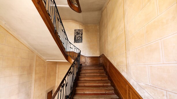 Un escalier avec des marches en bois et des rampes en métal noir, menant à un palier orné d’un élément décoratif mural.