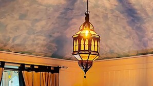 Une chambre à coucher avec un lit, un luminaire suspendu, une étagère fixée au mur ornée d’un objet décoratif et une fenêtre avec des rideaux.