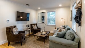 Una sala de estar con un sillón marrón, un sofá gris, una mesa de centro y un televisor de pantalla plana montado en la pared.