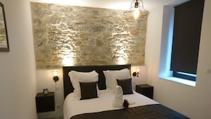 A bedroom with a stone wall, a bed with white and dark bedding, two bedside tables with lamps, and a window with a blind.