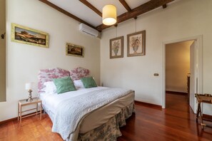 A bedroom with a wooden floor, a bed with floral bedding, a nightstand with a lamp, and two framed pictures on the wall.