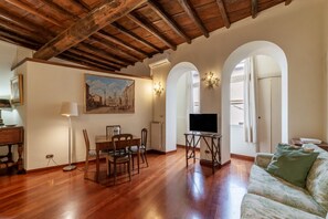 A spacious living room with wooden flooring, a dining table with chairs, a sofa, a TV on a stand, and a painting on the wall.